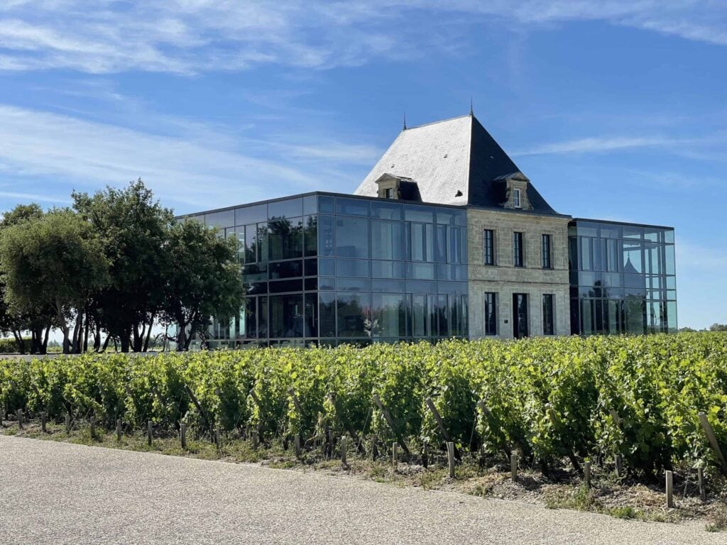 Château Pédesclaux in Pauillac, Grand Cru Classé des Médoc, ein modernes Gebäude mit Glasfassaden und einem zentralen Steinteil, umgeben von grünen Weinbergen unter blauem Himmel, in der Weinregion Bordeaux.