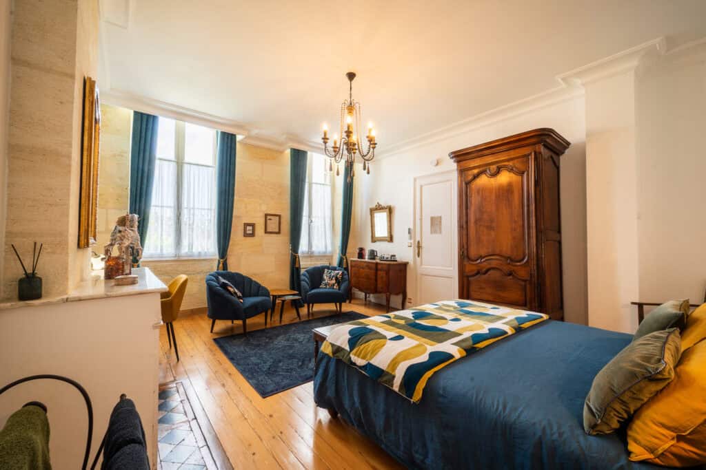 Vue intérieure de la Chambre Domeyne avec lit, fauteuils, armoire ancienne et décoration rétro au Château Papounan, chambre d’hôtes de charme à Saint-Estèphe, Médoc.